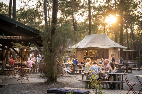 Zona de comedor al aire libre en Huttopia Lac de Carcans, Francia, con gente disfrutando entre pinos.