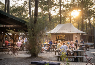 Udendørs spiseområde ved Huttopia Lac de Carcans feriepark i Frankrig, med gæster, træer og sollys.