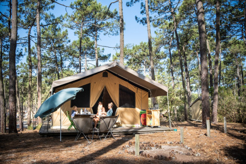 Familia sentada frente a una tienda safari en un bosque de pinos en Huttopia Lac de Carcans, Nouvelle-Aquitaine.
