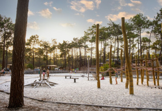 Kinder spielen auf einem naturbelassenen Spielplatz mit Sand, umgeben von Kiefern im Huttopia Lac de Carcans.