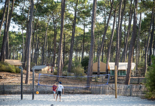 Children play volleyball on a sandy court among pine trees and tents at Huttopia Lac de Carcans, France.