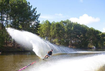 Wasserskifahrer gleitet durch einen Kanal bei Carcans, umgeben von grünen Bäumen und blauem Himmel.