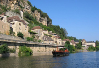 Boat on river by cliffside houses and greenery near Huttopia Sarlat holiday park in Nouvelle-Aquitaine, France.