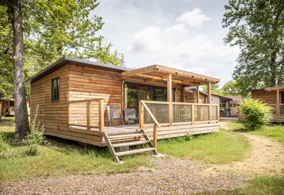 Cabaña de madera llamada Chalet Portland con porche, ubicada en zona boscosa y soleada, rodeada de árboles.
