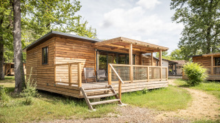 Cabaña de madera llamada Chalet Portland con porche, ubicada en zona boscosa y soleada, rodeada de árboles.