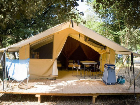 Safari Bungalow tent Classic on wooden deck, surrounded by trees, with chairs, table and camping gear in front.