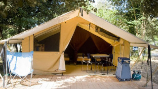Safari-Bungalowzelt Classic auf Holzplattform, mit Stühlen, Tisch und Campingausrüstung, umgeben von Bäumen.