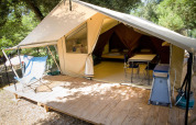 Tenda safari aperta su pedana di legno nel bosco, con sedie, tavolo e letti all'interno visibili.