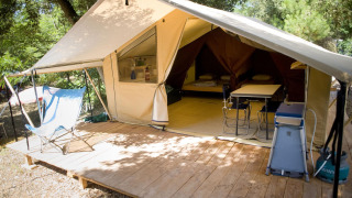 Tienda safari de lona abierta sobre plataforma de madera en el bosque, con sillas, mesa y camas dentro.