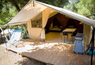 Tienda safari de lona abierta sobre plataforma de madera en el bosque, con sillas, mesa y camas dentro.