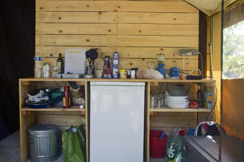 Küchenbereich im Safari-Zelt Toile&Bois Sweet mit Holzwand, Kühlschrank und Campingutensilien.
