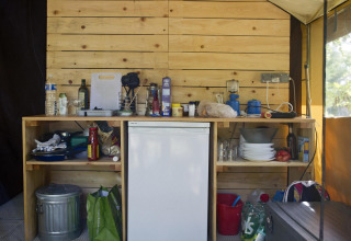 Küchenbereich im Safari-Zelt Toile&Bois Sweet mit Holzwand, Kühlschrank und Campingutensilien.