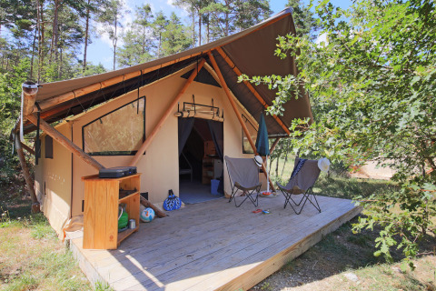 Safari tent Trappeur Tent with wooden deck, two chairs, umbrella and storage cabinet in nature setting.