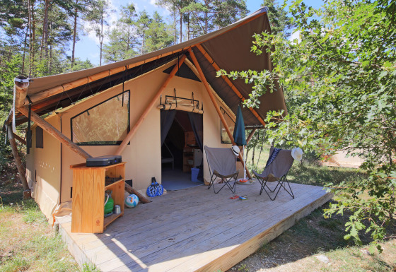 Tenda safari Trappeur Tent con terrazza in legno, due sedie, ombrellone e mobile in ambiente naturale.