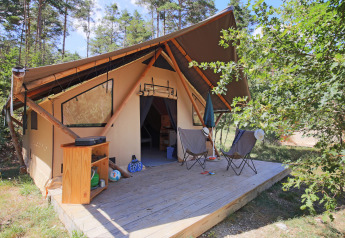 Safari tent Trappeur Tent with wooden deck, two chairs, umbrella and storage cabinet in nature setting.