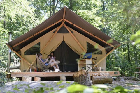 Two people relax in front of a Trappeur safari tent in the forest, surrounded by trees and nature.
