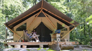 Dos personas se relajan frente a una tienda safari Trappeur en el bosque, rodeadas de árboles.