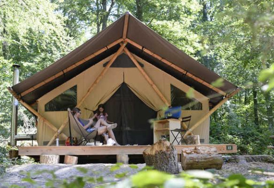 Dos personas se relajan frente a una tienda safari Trappeur en el bosque, rodeadas de árboles.