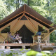 Dos personas se relajan frente a una tienda safari Trappeur en el bosque, rodeadas de árboles.