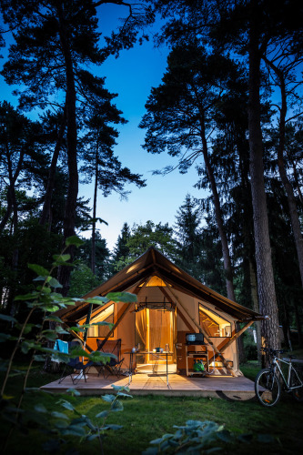 Illuminated Trappeur tent at Huttopia Baie du Mont St Michel, France, surrounded by tall pine trees.