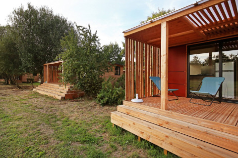Moderne houten chalet met overdekte veranda, twee stoelen en omgeven door gras en bomen.