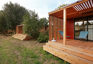 Modern wooden cabin with a covered deck, two chairs, and trees in the background on a grassy lawn.