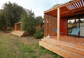 Cabaña de madera moderna con terraza cubierta, dos sillas y árboles y césped verde en el entorno.