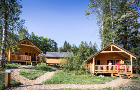 Cabañas de madera en Chalet Montana, Huttopia Bozel en Vanoise, Francia, rodeadas de vegetación y árboles altos bajo el sol.