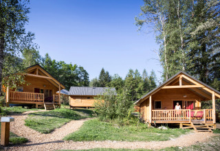 Cabañas de madera en Chalet Montana, Huttopia Bozel en Vanoise, Francia, rodeadas de vegetación y árboles altos bajo el sol.