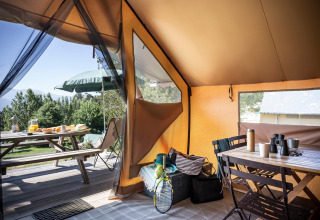 Vista interna di una tenda safari con tavolo, sedie e borse, verso un’area picnic verde all’esterno.