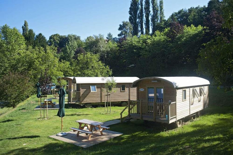 Foto di due cabine in legno tra alberi verdi e tavoli da picnic con ombrelloni, in una giornata di sole.