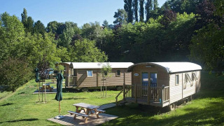 Photo of two wooden cabins in a lush, green setting with picnic tables and umbrellas on a sunny day.