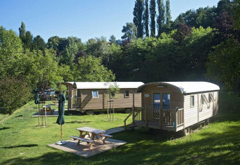 Photo of two wooden cabins in a lush, green setting with picnic tables and umbrellas on a sunny day.