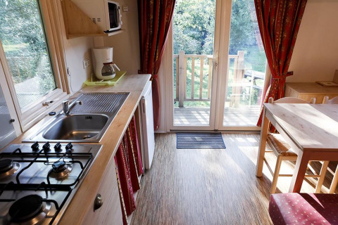 Knusse keuken in een houten hut met gaskookplaat, koffiemachine, eethoek en zicht op groene tuin via glasdeur.