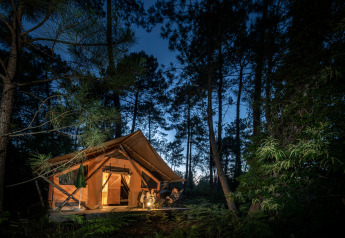 Illuminated Trappeur safari tent in a forest at night with people relaxing by the tent entrance.
