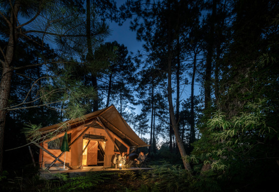 Tente safari Trappeur éclairée dans la forêt la nuit, avec des personnes posées devant l'entrée.