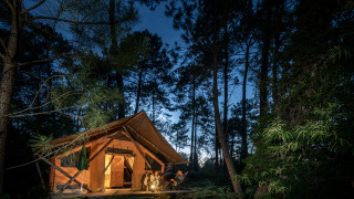 Illuminated Trappeur safari tent in a forest at night with people relaxing by the tent entrance.