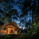 Tienda safari Trappeur iluminada en un bosque por la noche con personas sentadas en la entrada.