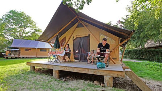 Family relaxing on the porch of a Trappeur II safari tent, surrounded by greenery and summer sun.