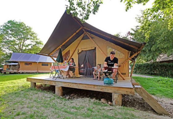 Family relaxing on the porch of a Trappeur II safari tent, surrounded by greenery and summer sun.