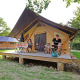 Familia relajándose en el porche de una tienda safari Trappeur II, rodeada de naturaleza verde.