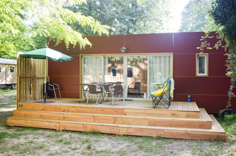 Cabaña moderna con terraza de madera, muebles de exterior, sombrilla verde y rodeada de árboles.
