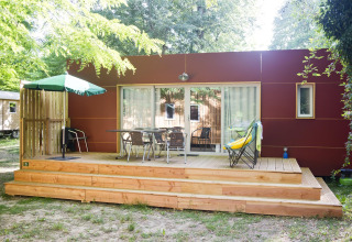 Cabaña moderna con terraza de madera, muebles de exterior, sombrilla verde y rodeada de árboles.
