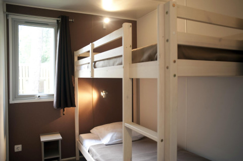 A simple cabin room with wooden bunk beds, a window with curtains, and a small bedside table in the corner.