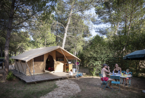 Familie frühstückt an einem Tisch vor einem Safarizelt im Wald bei Huttopia Fontvieille, Frankreich.