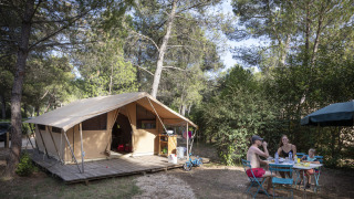 Familie, der nyder morgenmad ved et bord foran en safaritelthytte i skovene ved Huttopia Fontvieille, Frankrig.