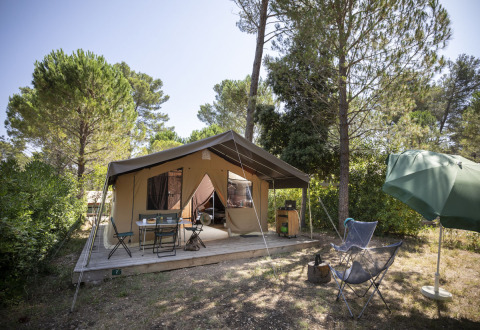 Tienda safari Toile&Bois Sweet en camping forestal, con terraza, sillas exteriores y sombrilla verde.