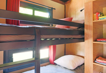 Glamping-Unterkunft mit Etagenbetten, Holzregalen und Fenstern mit roten Vorhängen im Sonnenlicht.