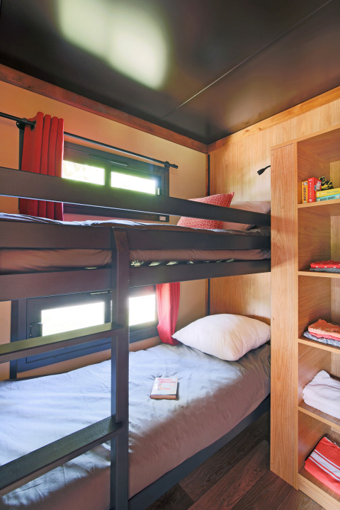 Habitación de glamping con literas, estantería de madera y ventanas con cortinas rojas en plena luz del día.
