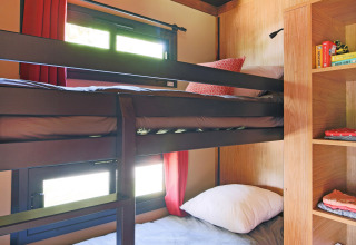 Glamping room with bunk beds, wood shelving, storage cubbies, and windows with red curtains in daylight.